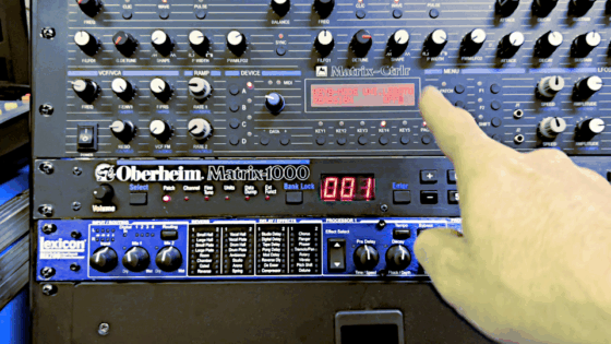 An Oberheim Matrix-1000 in a rack, below the Alpes Machines Ctrlr with loads of knobs, switched on, and a Lexicon MX200 reverb/delay rack effect. Finger pointing at the display of the Alpes controller showing the Rob/Reassign setting which does not work pre-V1.21.